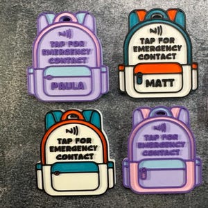 May include: Four backpack-shaped tags featuring a raised NFC symbol and the text TAP FOR EMERGENCY CONTACT. Two tags are purple with the name PAULA, while the other two are white with orange and teal accents, one labeled with the name MATT.