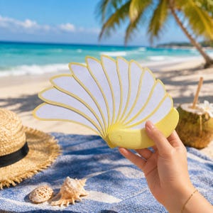 May include: A handheld folding fan with a yellow handle and frame, featuring multiple white, petal-shaped blades with yellow edges. The fan is held in a hand against a backdrop of a sandy beach, blue ocean, a straw hat, and a coconut with a straw.