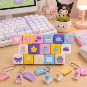 May include: A collection of square mechanical keyboard keycaps featuring pastel-coloured icons including hearts, flowers, stars, and food items. Below the stacked keycaps are several small, clear mechanical switch keychains with attached tags labelled with words like Click, Cute, and Happy.