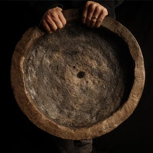 May include: A large, circular, dark brown wooden bowl with a thick, uneven rim and a rough, textured interior surface. The center of the bowl features a small, distinct circular hole, and the wood grain shows natural variations in color and depth.