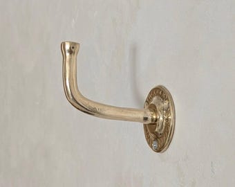 Puede incluir: Un gancho de pared de color latón con un brazo curvo y una base circular decorativa. La base está adornada con un patrón en relieve. El gancho está montado en una pared de color claro.