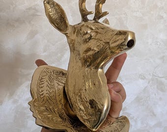 Puede incluir: Una escultura de cabeza de ciervo de latón con cornamenta detallada y una base decorativa. La escultura se sostiene en una mano, mostrando su acabado pulido y su intrincado diseño. La cabeza de ciervo es un objeto decorativo.