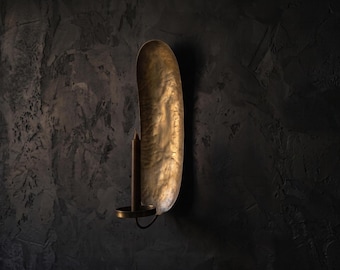 Puede incluir: Un candelabro de pared que presenta una placa trasera de latón alta y ovalada con un acabado martillado y texturizado. Una copa cilíndrica para velas está unida a la sección inferior de la placa, diseñada para sostener una vela cónica.