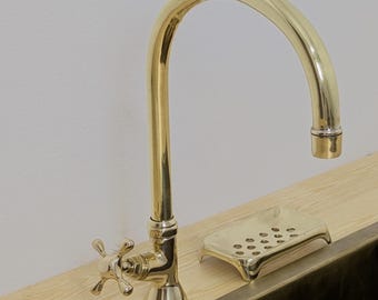 Puede incluir: Un grifo dorado con un caño curvo y manijas en forma de cruz está montado sobre una superficie de madera clara. Una jabonera a juego se encuentra junto al grifo. El fregadero es de metal oscuro.