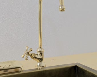 Puede incluir: Un grifo dorado con un caño curvo y manijas en forma de cruz. El grifo está montado sobre una superficie de madera de color claro, con un fregadero de color oscuro debajo. El fondo es una pared lisa de color claro.