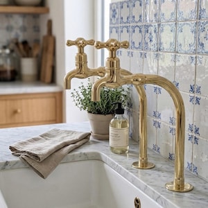 Può includere: Un lavello da cucina con due rubinetti dorati. Un piccolo vaso di piante e una bottiglia di sapone sono sul piano di lavoro in marmo. Un asciugamano di lino beige piegato è appoggiato sul bancone, con piastrelle a motivi blu e bianchi sullo sfondo.