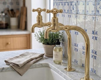 May include: A kitchen sink with a marble countertop features two gold-colored faucets. A small potted plant and a bottle of soap sit near the faucets. A folded beige linen towel rests on the counter, with blue and white patterned tiles in the background.