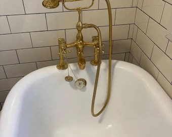 Puede incluir: Un conjunto de grifería para bañera en latón pulido que incluye un cabezal de ducha de mano con manguera flexible, dos mandos en cruz y un caño central. El accesorio está montado en una bañera de porcelana blanca con desagüe de latón.