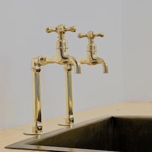 May include: Two gold-colored faucets with cross-shaped handles are mounted on a light-colored wooden surface. The faucets are positioned above a dark-colored rectangular sink. The design is classic and elegant, suitable for a kitchen or bathroom.