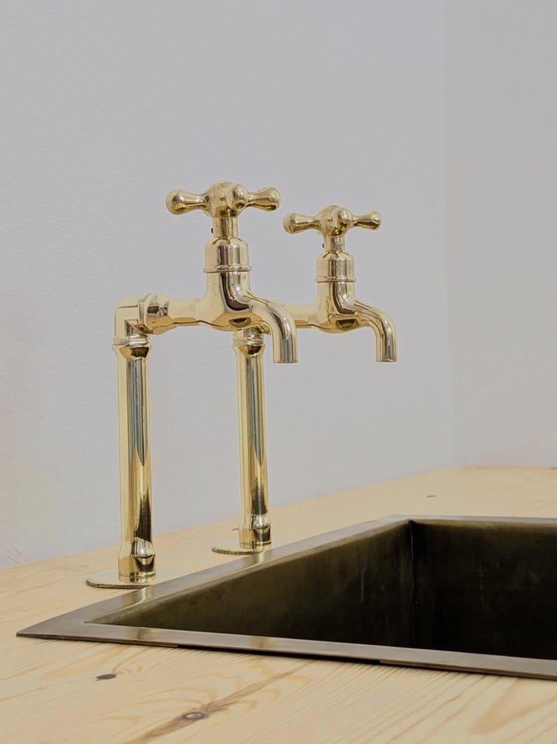 May include: Two gold-colored faucets with cross-shaped handles are mounted on a light-colored wooden surface. The faucets are positioned above a dark-colored rectangular sink. The design is classic and elegant, suitable for a kitchen or bathroom.