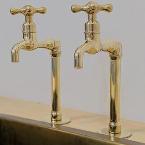May include: Two gold-colored faucets with cross-shaped handles are mounted on a wooden surface. The faucets have a classic design with curved spouts and vertical pipes. The background is a plain white wall.