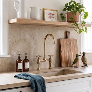 Könnte beinhalten: Küchenszene mit Messingarmatur, Holzschneidebrett und dekorativen Elementen. Zwei weiße Krüge und ein gerahmter Fischdruck stehen auf einem Holzregal. Zwei braune Seifenspender und ein blaues Tuch befinden sich auf der Arbeitsplatte. Zwei Holzfiguren von Vögeln sind ebenfalls vorhanden.