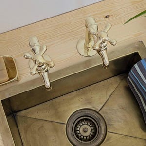 May include: A brass-toned sink with two matching faucets. The faucets have cross-shaped handles. The sink is set into a light-colored wooden countertop. A small soap dish and a striped towel are visible.
