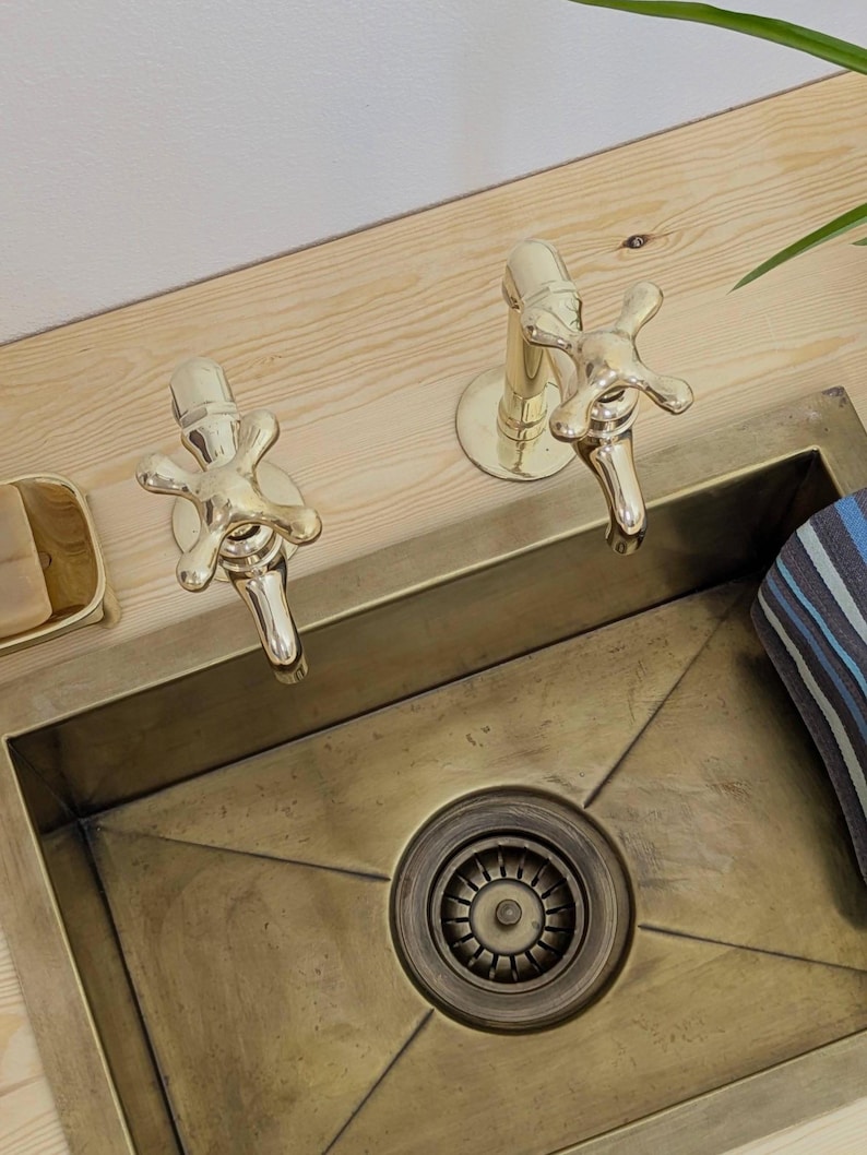 May include: A brass-toned sink with two matching faucets. The faucets have cross-shaped handles. The sink is set into a light-colored wooden countertop. A small soap dish and a striped towel are visible.