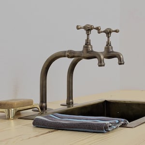 May include: Two antique brass taps with curved spouts are mounted on a light wood surface. A folded striped towel in shades of blue, grey, and white rests near a dark metal sink. A bar of soap sits in a brass soap dish.