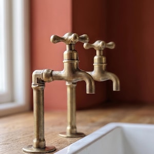 May include: Two antique-style brass faucets with cross-shaped handles and cylindrical spouts. The faucets feature a weathered metallic finish and are mounted on a wooden surface next to a white ceramic sink basin, showcasing a classic design.