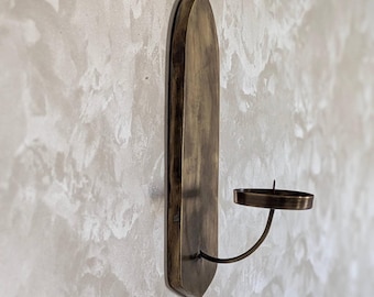 Puede incluir: Un portavelas de pared de latón. Presenta una placa trasera ovalada y alargada y un brazo curvado que sostiene una copa para velas circular. El diseño es sencillo y elegante, adecuado para varios estilos de interior. Medidas en centímetros.