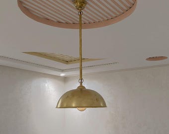 Puede incluir: Una lámpara colgante de latón con una pantalla en forma de cúpula, suspendida por una varilla delgada desde un soporte de techo circular con un patrón de rayas rosas y blancas. La superficie metálica de la lámpara tiene un acabado pulido.