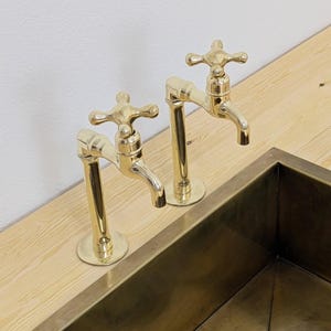 May include: Two polished brass faucets with cross-shaped handles are mounted on a light wood surface. A matching brass sink is partially visible below. The faucets have a classic design, adding a touch of elegance to the kitchen or bathroom.
