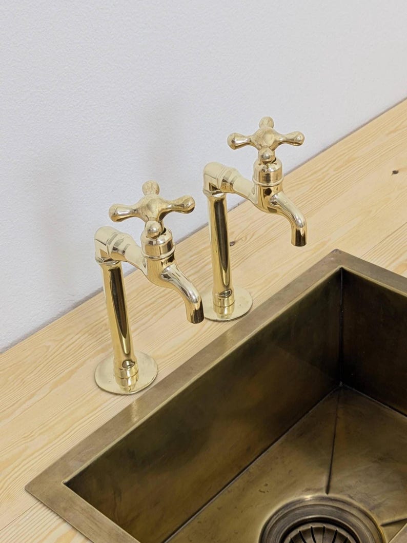 May include: Two polished brass faucets with cross-shaped handles are mounted on a light wood surface. A matching brass sink is partially visible below. The faucets have a classic design, adding a touch of elegance to the kitchen or bathroom.