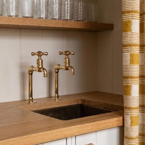 May include: A kitchen sink area with two brass faucets, a dark sink basin, and a wooden countertop. Clear drinking glasses sit on a wooden shelf above. A yellow and white striped curtain is on the right.