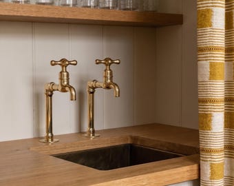 May include: A kitchen sink area with two brass faucets, a dark sink basin, and a wooden countertop. Clear drinking glasses sit on a wooden shelf above. A yellow and white striped curtain is on the right.