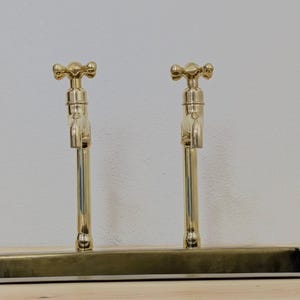 May include: Two gold-colored faucets with cross-shaped handles are mounted on a rectangular base. The faucets have a classic design and are set against a neutral background. The fixtures are made of brass.