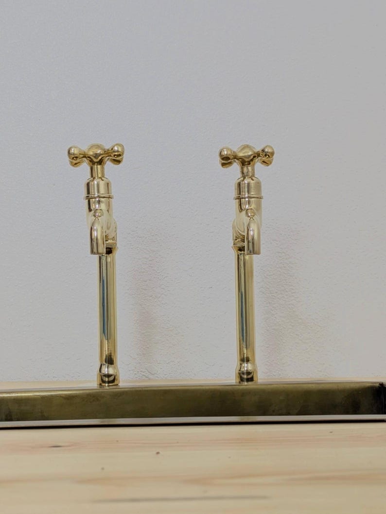 May include: Two gold-colored faucets with cross-shaped handles are mounted on a rectangular base. The faucets have a classic design and are set against a neutral background. The fixtures are made of brass.