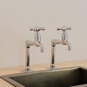 May include: Two polished chrome faucets with cross-handle knobs are mounted on a light wood surface behind a rectangular, dark metallic sink basin. The faucets feature a vertical pipe design with a curved spout and a classic four-point handle.