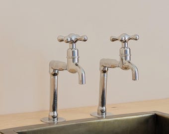 Puede incluir: Dos grifos de cromo pulido con mandos en cruz están montados sobre una superficie de madera clara detrás de un fregadero rectangular de metal oscuro. Los grifos presentan un diseño de tubo vertical con caño curvo y mando clásico.