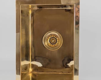 Puede incluir: Un fregadero de cocina rectangular de color dorado con un desagüe. El fregadero tiene un acabado pulido y un borde elevado. El desagüe es circular con un patrón decorativo. El fregadero es de metal y tiene un diseño moderno.