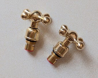 Puede incluir: Dos manijas de grifo de metal en tono dorado pulido con un diseño en forma de cruz y extremos redondeados. Cada manija tiene una base cilíndrica con una junta tórica de goma negra y una pequeña inserción circular roja en la punta inferior.