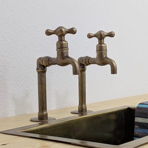 Puede incluir: Dos grifos antiguos de latón con manijas en cruz están montados sobre un fregadero rectangular. Los grifos tienen una estética vintage, con un acabado cálido y envejecido. El fregadero es de metal oscuro y está empotrado en una encimera de madera clara.