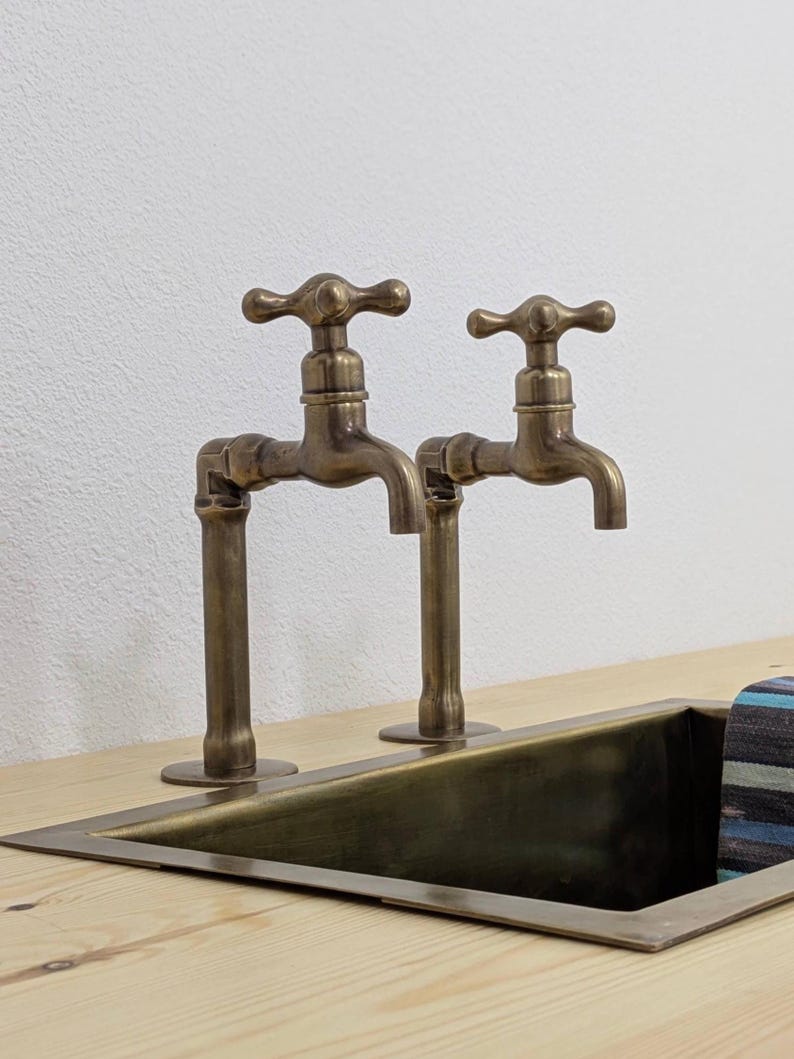 Puede incluir: Dos grifos de lat&oacute;n de estilo antiguo con mandos en cruz, montados sobre una encimera de madera clara junto a un fregadero de metal rectangular. Los grifos presentan un dise&ntilde;o de tuber&iacute;a vertical con un acabado met&aacute;lico oscuro.