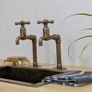 Puede incluir: Dos grifos de lat&oacute;n antiguo con mandos en cruz montados sobre una encimera de madera clara. Un lavabo rectangular de metal se sit&uacute;a bajo los grifos, acompa&ntilde;ado de una peque&ntilde;a jabonera y un pa&ntilde;o plegado de rayas azules y grises.