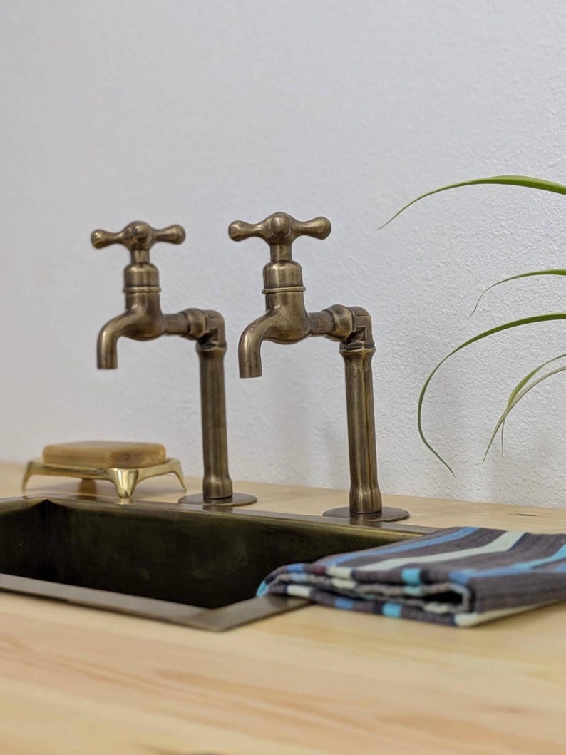 Puede incluir: Dos grifos de lat&oacute;n antiguo con mandos en cruz montados sobre una encimera de madera clara. Un lavabo rectangular de metal se sit&uacute;a bajo los grifos, acompa&ntilde;ado de una peque&ntilde;a jabonera y un pa&ntilde;o plegado de rayas azules y grises.