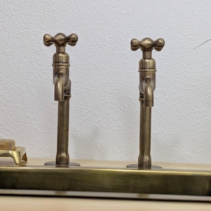 May include: Two antique brass taps with cross-shaped handles. The taps are mounted on a rectangular base, displaying a vintage design. The warm brass colour adds a classic touch to the bathroom fixtures.