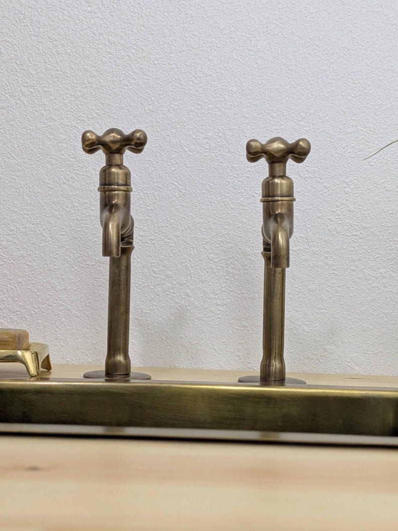Puede incluir: Dos grifos con acabado en lat&oacute;n antiguo y manijas en forma de cruz montados sobre una barra horizontal. Los grifos presentan un cuerpo cil&iacute;ndrico vertical y un ca&ntilde;o orientado hacia abajo. Un peque&ntilde;o jabonero de lat&oacute;n es visible a la izquierda.