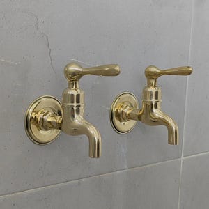 Può includere: Due rubinetti color oro montati su una parete piastrellata grigio chiaro. Ogni rubinetto ha un beccuccio curvo, una base rotonda e una maniglia a leva. I rubinetti sono posizionati uno accanto all'altro, suggerendo un ambiente bagno o cucina.