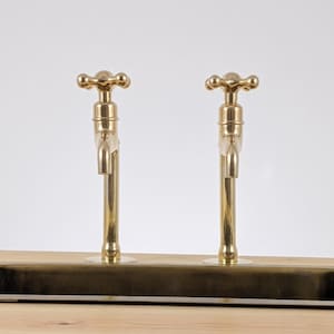 May include: Two brass faucets with cross-shaped handles are mounted on a wooden surface. The faucets have a classic design with a polished finish. The background is a plain white, highlighting the fixtures.