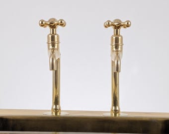 May include: Two brass faucets with cross-shaped handles are mounted on a wooden surface. The faucets have a classic design with a polished finish. The background is a plain white, highlighting the fixtures.