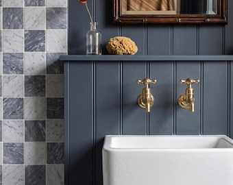 Puede incluir: Un lavabo de pared de cerámica blanca con dos grifos de latón con manijas en cruz montados sobre una pared de paneles verticales gris oscuro. Un jarrón de vidrio transparente con una flor roja y una esponja natural descansan en el estante.
