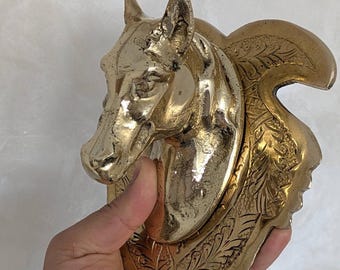 Puede incluir: Una escultura de cabeza de caballo dorada con detalles precisos. La escultura tiene una superficie texturizada y se sostiene en una mano. La cabeza de caballo está montada en una placa decorativa con un diseño de hojas ornamentado.