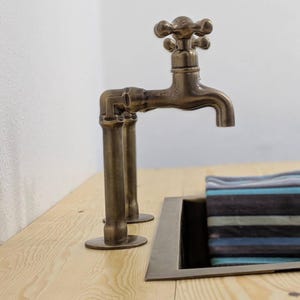 Puede incluir: Un grifo de metal en tono bronce con dise&ntilde;o de manija en cruz, que presenta una estructura similar a una tuber&iacute;a montada sobre una superficie de madera clara junto a un lavabo rectangular con una tela a rayas azules y grises.