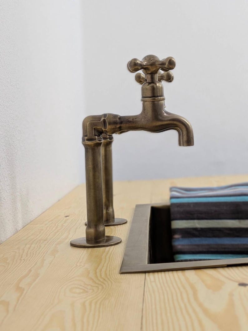 Puede incluir: Un grifo de metal en tono bronce con dise&ntilde;o de manija en cruz, que presenta una estructura similar a una tuber&iacute;a montada sobre una superficie de madera clara junto a un lavabo rectangular con una tela a rayas azules y grises.