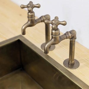 Puede incluir: Dos grifos de lat&oacute;n antiguo con manijas en forma de cruz montados sobre una superficie de madera clara encima de un fregadero rectangular de lat&oacute;n. Los grifos presentan un dise&ntilde;o de tuber&iacute;a con un ca&ntilde;o curvo y un acabado met&aacute;lico desgastado.