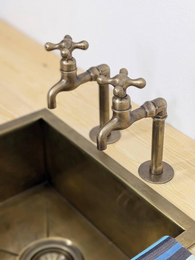 Puede incluir: Dos grifos de lat&oacute;n antiguo con manijas en forma de cruz montados sobre una superficie de madera clara encima de un fregadero rectangular de lat&oacute;n. Los grifos presentan un dise&ntilde;o de tuber&iacute;a con un ca&ntilde;o curvo y un acabado met&aacute;lico desgastado.