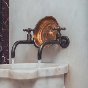 May include: An antique bronze tap with two curved spouts, mounted on a white marble wall. A decorative copper plate with an etched design is centered above the tap. Water flows from the spouts into a white marble sink.