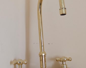 Puede incluir: Un grifo de cocina de latón pulido con un caño de arco alto y dos manijas en forma de cruz. El accesorio tiene un tubo base horizontal que conecta las dos manijas con el caño central, mostrando un acabado dorado metálico en toda la unidad.