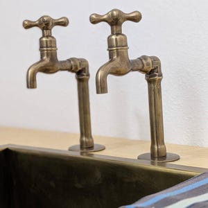 Puede incluir: Dos grifos de lat&oacute;n de estilo antiguo con mandos en cruz montados sobre una superficie de madera clara. Los grifos presentan un acabado met&aacute;lico desgastado y est&aacute;n situados detr&aacute;s de un lavabo rectangular de lat&oacute;n con un pa&ntilde;o a rayas.